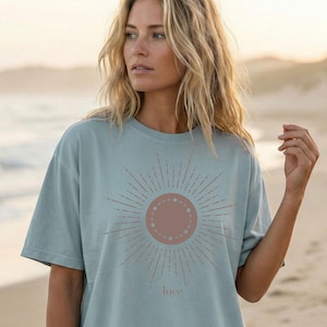 Könnte beinhalten: Hellblaues T-Shirt mit einem rosafarbenen Sonnenmuster und dem Wort "luce" darunter. Das T-Shirt hat kurze Ärmel und eine lockere Passform. Geeignet für Freizeitkleidung.
