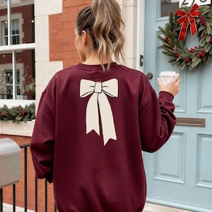 May include: Burgundy sweatshirt with a large white bow graphic on the back. The sweatshirt is a solid color and has a crew neck. The person is holding a white coffee cup. The background includes a wreath and a door.
