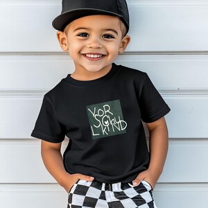 May include: A young child wearing a black t-shirt with the words "Your Lucky Kind" in white script inside a green square. The child is also wearing a black baseball cap and checkered pants. The child is smiling.