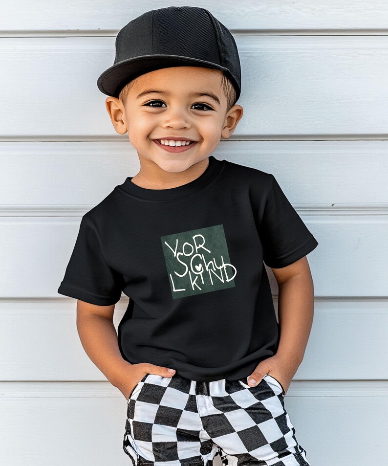 May include: A young child wearing a black t-shirt with the words "Your Lucky Kind" in white script inside a green square. The child is also wearing a black baseball cap and checkered pants. The child is smiling.