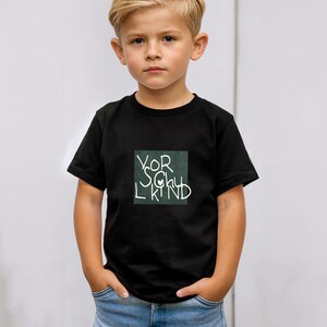 May include: A black t-shirt with the words "VOR SCHU L KIND" in white script inside a green square. The child is wearing blue jeans and has their hands in their pockets.
