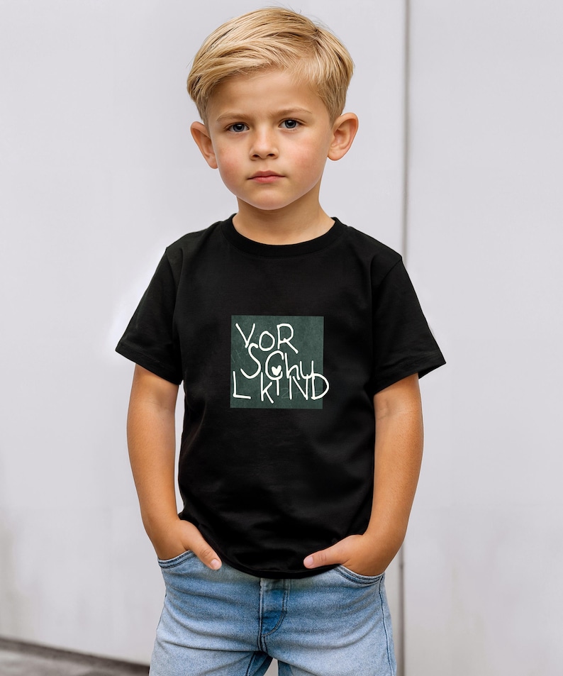 May include: A black t-shirt with the words "VOR SCHU L KIND" in white script inside a green square. The child is wearing blue jeans and has their hands in their pockets.