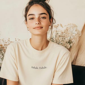 May include: A cream-colored t-shirt with the words "Inhale. Exhale." printed in black. The shirt is worn by a person with brown hair pulled up in a bun. The background includes flowers and a neutral-toned wall.