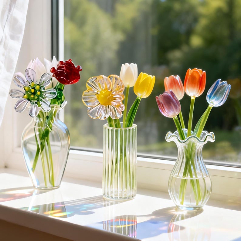 May include: Three glass vases with colorful glass flowers sit on a windowsill. The flowers include a daisy, tulips in shades of red, yellow, orange, purple, and blue. The vases are clear glass, and the sunlight casts colorful shadows.