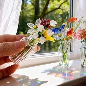 May include: A clear glass vase with a bouquet of colorful glass flowers, including white, yellow, red, and blue blooms. The vase is held in a hand near a window with other glass flower arrangements. The flowers are detailed and appear delicate.