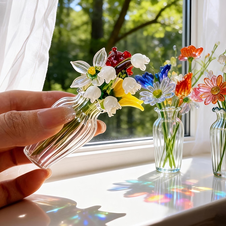 May include: A clear glass vase with a bouquet of colorful glass flowers, including white, yellow, red, and blue blooms. The vase is held in a hand near a window with other glass flower arrangements. The flowers are detailed and appear delicate.