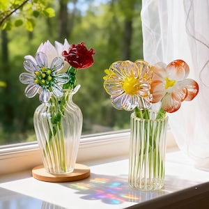 May include: Two glass vases with colorful glass flowers sit on a windowsill. One vase holds a white, red, and clear flower arrangement. The other vase holds a yellow and orange flower arrangement. The vases are clear and have a ribbed design.