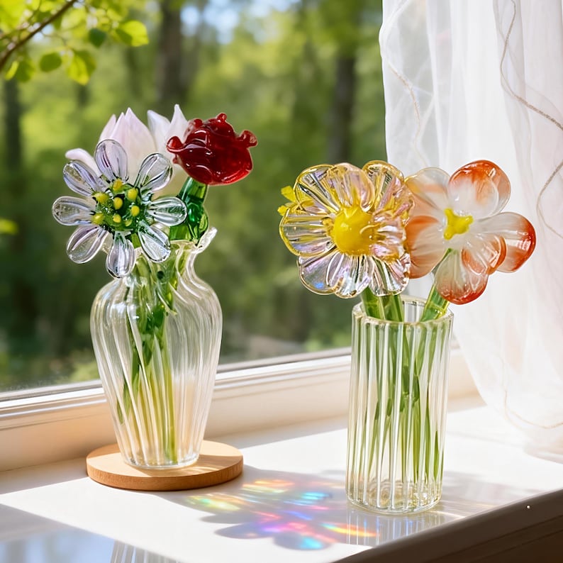 May include: Two glass vases with colorful glass flowers sit on a windowsill. One vase holds a white, red, and clear flower arrangement. The other vase holds a yellow and orange flower arrangement. The vases are clear and have a ribbed design.