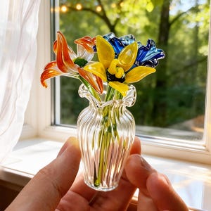 May include: A small glass vase with a ruffled rim holds colorful glass flowers. The flowers are orange, yellow, and blue. The vase is clear and has vertical ridges. The arrangement is set near a window.