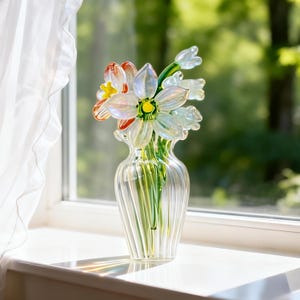 May include: A clear glass vase with a fluted design holds a bouquet of glass flowers. The flowers include white, orange, and yellow petals with green stems. The vase sits on a white windowsill with a sheer white curtain in the background.