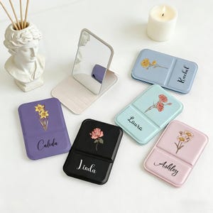 May include: A collection of personalized compact mirrors in various colors, each adorned with a floral design and a name. The colors include purple, black, blue, and pink. A silver-framed mirror is also present, along with a lit candle and a bust.