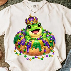 May include: White t-shirt with a cartoon frog wearing a crown and sitting on a decorated pastry. The frog is green with a smiling face. The pastry is decorated with purple, green, and gold beads. The shirt has the words "Comfort Colors" printed on it.