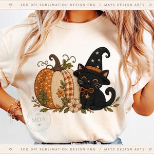 May include: A cream-colored t-shirt featuring a graphic of a black cat wearing a witch hat with stars, sitting next to a large pumpkin with orange and white patterns, decorated with small flowers and green leaves, with the text MDA visible.