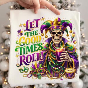 Könnte beinhalten: Weißes T-Shirt mit einem farbenfrohen Mardi-Gras-Design. Die Grafik zeigt ein Skelett mit Narrenhut, das ein Getränk hält, mit dem Text "Let the Good Times Roll!" in Lila, Grün und Gold.