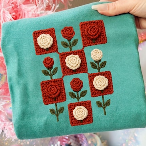 Könnte beinhalten: Ein türkisblaues T-Shirt mit einem Häkeldesign aus roten Quadraten mit cremefarbenen und roten Rosen. Die Rosen sind durch grüne Stiele und Blätter verbunden. Das Design ist auf dem T-Shirt zentriert.