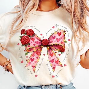 Op de afbeelding: Crèmekleurige sweatshirt met een grote roze strik versierd met rode harten en rozen. De strik is versierd met lichtslingers en omgeven door de zinnen "You are my sunshine" en "You are a treasure" in een handschrift.