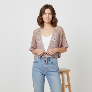 Pode incluir: Um cardigan aberto cor de malva com mangas curtas e fecho de laço. O cardigan tem um design delicado de malha aberta. A modelo está vestindo um top branco e jeans azuis. Um banco de madeira está no fundo.