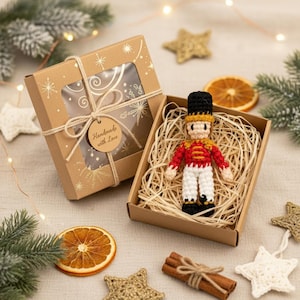 Könnte beinhalten: Gehäkelte Nussknacker-Puppe in einer Geschenkbox. Die Puppe trägt eine rote Jacke, eine weiße Hose und einen schwarzen Hut. Die Geschenkbox ist braun mit einem transparenten Fenster und einem Etikett "Handmade with Love".