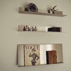 May include: Three stainless steel floating shelves mounted on a wall. The top shelf holds a purple geode and a bronze bird sculpture. The middle shelf displays decorative jars and books. The bottom shelf features a vintage Vogue magazine and a leather-bound book.