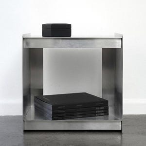 May include: A modern, brushed metal side table with a rectangular frame. A small, black, hexagonal box sits on top. Inside, a shelf holds a stack of black books. The table is minimalist and industrial in style.