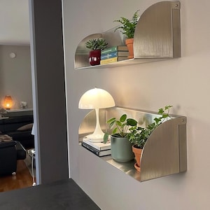 Puede incluir: Dos estanterías de pared de metal cepillado con diseño semicircular. Cada estantería contiene plantas en macetas, libros y una lámpara blanca. Las estanterías están montadas en una pared gris claro, creando una estética moderna y minimalista.