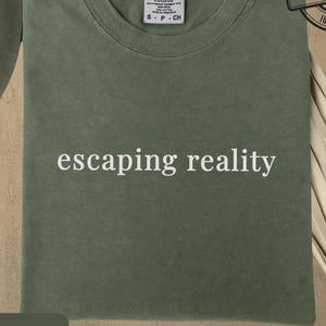 May include: A moss green t-shirt with the phrase "escaping reality" printed in white. The shirt is made of 100% cotton and features a Comfort Colors label. The shirt is a small size.