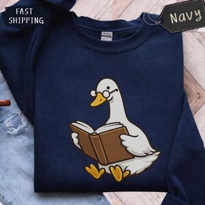 Puede incluir: Sudadera azul marino con un dibujo de un ganso de dibujos animados con gafas y leyendo un libro. El ganso es blanco con pico y patas amarillas. La sudadera tiene la palabra "Navy" en una etiqueta y las palabras "Fast Shipping".
