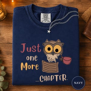 May include: A navy blue t-shirt featuring a graphic of an owl holding a book and a mug. The text reads "Just one More ...CHAPTER." The shirt has a Comfort Colors brand label at the neck and a silver-toned star charm necklace resting on the collar.