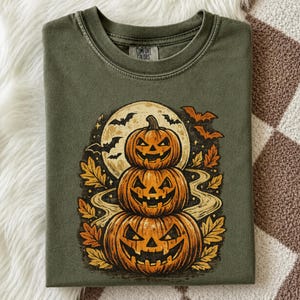 May include: An olive green t-shirt featuring a graphic print of three stacked jack-o'-lanterns in front of a full moon. The design includes flying bats, swirling paths, and autumn leaves in shades of orange and yellow. The shirt has a crew neckline.
