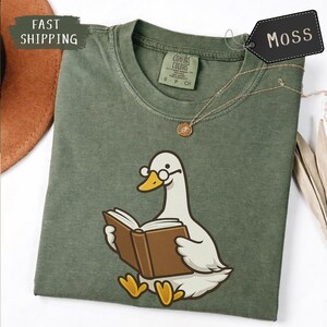 Puede incluir: Una camiseta verde musgo con un pato de dibujos animados con gafas leyendo un libro. La camiseta tiene cuello redondo y mangas cortas. Se ve un collar dorado con un colgante. La etiqueta dice "MOSS".