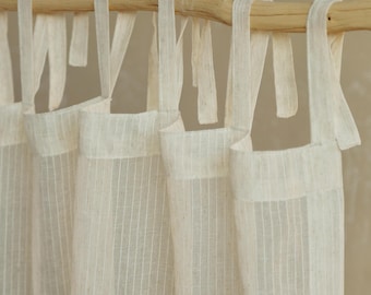 May include: Off-white linen curtains with vertical stripes, hanging from a natural wooden rod. The curtains are tied with matching fabric ties, creating a soft, textured look. The neutral color palette suggests a minimalist or farmhouse style.