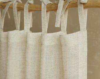 May include: Off-white linen curtains with tie-top closures, hanging from a natural wooden rod. The curtains have a textured weave and are slightly gathered, creating soft folds. The neutral color and simple design suggest a minimalist or farmhouse style.