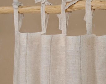 May include: Beige linen curtains with vertical stripes, hanging from a natural wooden rod. The curtains are tied with fabric loops. The fabric has a textured appearance, suggesting a natural fiber.