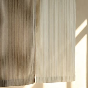 May include: Close-up of two fabric panels with vertical ribbed texture. One panel is a light beige colour, and the other is a darker tan. The fabric is lit by natural light, creating shadows and highlighting the texture.