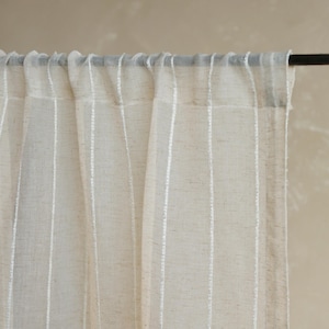 May include: Beige linen curtain with vertical white stripes. The curtain is hanging from a dark metal rod. The fabric appears semi-sheer, allowing light to filter through. The overall aesthetic is simple and natural.