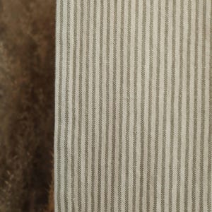 Puede incluir: Primer plano de una tela con rayas verticales. Las rayas son de color verde apagado sobre un fondo crema. La tela tiene un aspecto texturizado, lo que sugiere que puede ser lino o un material similar. La estética general es simple y clásica.