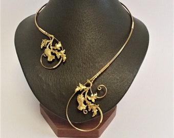 May include: A gold-colored necklace with a leaf and vine design, displayed on a black mannequin. The necklace features a curved, open design with intricate leaf details and a circular element. The jewelry is presented against a white background.
