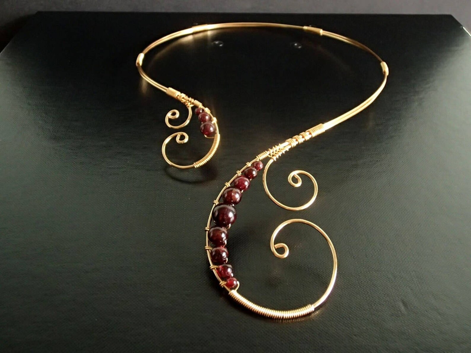 Handmade gold collar necklace with asymmetrical garnet bead swirl design on black background.