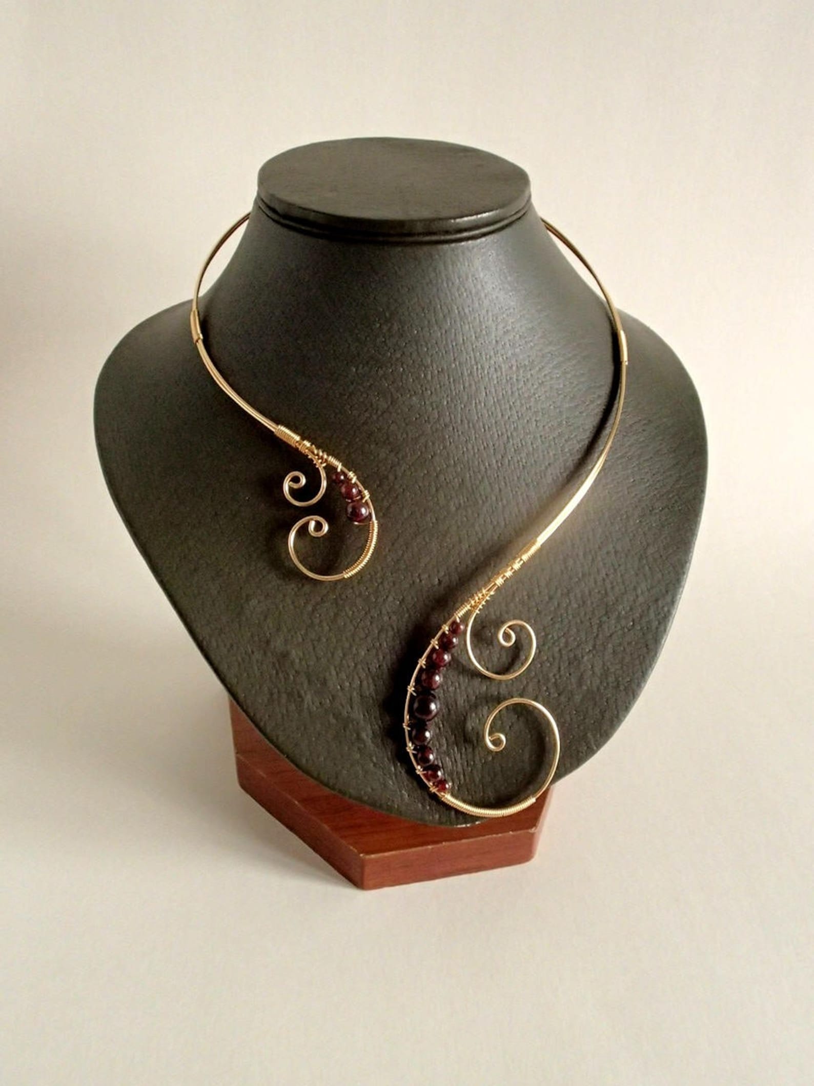 Gold wire collar necklace with swirling asymmetrical design and graduated garnet beads on display stand.