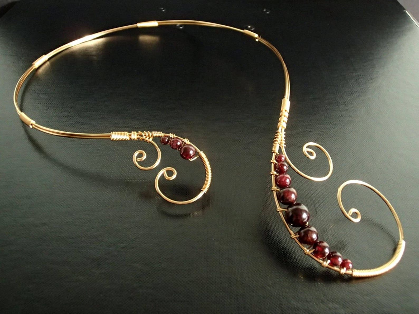 Asymmetric open gold collar necklace with red garnet beads arranged in a swirling wire design.