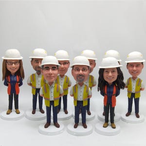 以下が含まれることがあります： 建設作業員の首振り人形のコレクション。各フィギュアは白いヘルメット、安全ベスト、作業服を着用しています。フィギュアは白い円形の台座にあり、詳細な顔の特徴があります。