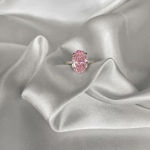 May include: A pink oval gemstone ring with a gold band, set against white satin. The ring features a large, pink central stone and a band embellished with smaller, clear stones. The satin fabric is draped, creating a luxurious background.