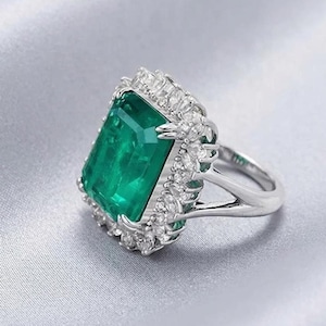 May include: A silver ring featuring a large, rectangular emerald-cut green gemstone. The emerald is surrounded by a halo of smaller, clear gemstones. The ring's band is a polished silver color.