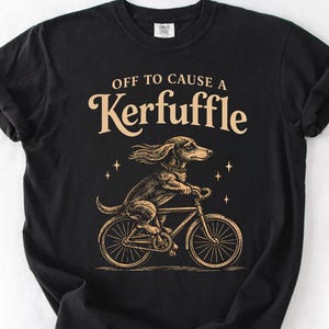 May include: Black t-shirt with a graphic of a dachshund riding a bicycle. The text "OFF TO CAUSE A Kerfuffle" is arched above the dog. The dog and bicycle are in a sepia tone.