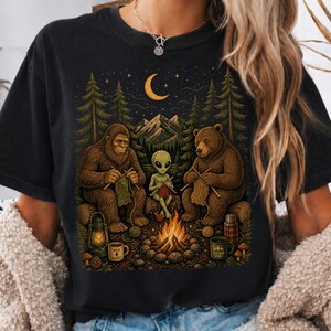 May include: Black t-shirt with a whimsical graphic of a Bigfoot, a bear, and an alien knitting around a campfire under a crescent moon. The words "Confort colors" are printed at the top left.