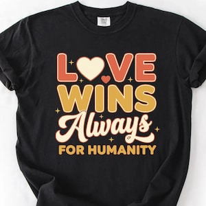 Può includere: T-shirt nero con la scritta "LOVE WINS Always FOR HUMANITY" in un font retrò. Il design presenta un cuore e stelle nei toni del rosso, giallo e bianco. La maglietta è realizzata in materiale morbido.