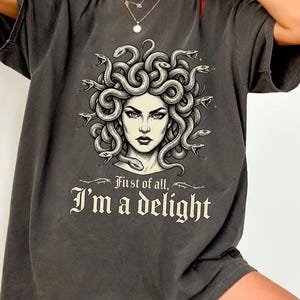 Könnte beinhalten: Ein anthrazitfarbenes Oversize-T-Shirt mit einer grafischen Illustration von Medusa mit Schlangen als Haaren. Unter dem Bild steht der Text "First of all, I'm a delight" in einer dekorativen, gotischen Schriftart. Das T-Shirt hat kurze Ärmel.