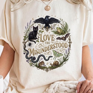 Könnte beinhalten: Ein cremefarbenes Kurzarm-T-Shirt mit einer kreisförmigen Grafik und dem Text LOVE THE MISUNDERSTOOD. Das Design zeigt einen schwarzen Raben, eine schwarze Katze, einen Frosch, eine Fledermaus, einen Nachtfalter, eine Schnecke, eine Maus und eine Spinne.