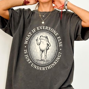 May include: A charcoal grey t-shirt featuring a circular graphic of a standing frog with one finger raised. The text surrounding the frog reads, "WHAT IF EVERYONE ELSE IS JUST UNDERTHINKING?" The design is rendered in a vintage, etched illustration style.