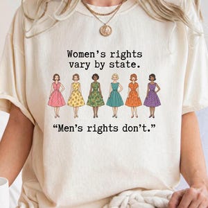 May include: An ivory short-sleeve t-shirt featuring a graphic of seven diverse figures in colorful dresses. The text reads: Women's rights vary by state. Men's rights don't. The shirt has a crew neckline and a relaxed fit, with the brand name Comfort Colors visible.